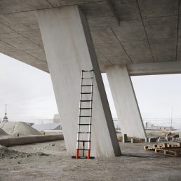 Een RMBO® Professionele telescopische ladder van 3,2 m staat tegen een schuine betonnen pilaar op een bouwplaats onder een nog niet voltooid viaduct, met bouwmaterialen en stadsgebouwen op de achtergrond.